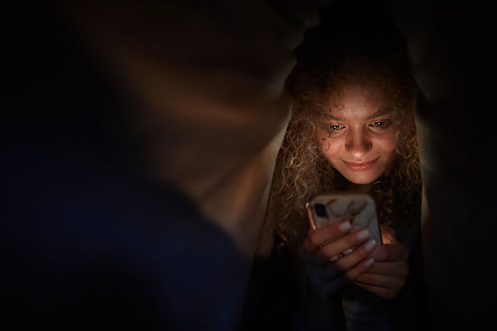 Young woman with curly hair smiling while looking at her phone in dim light, relating to faked twin brother confession. Young woman with curly hair smiling while looking at her phone in dim light, relating to faked twin brother confession.