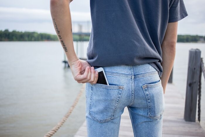 Person standing on a dock near water, secretly putting a phone into the back pocket of their jeans, capturing a candid moment.