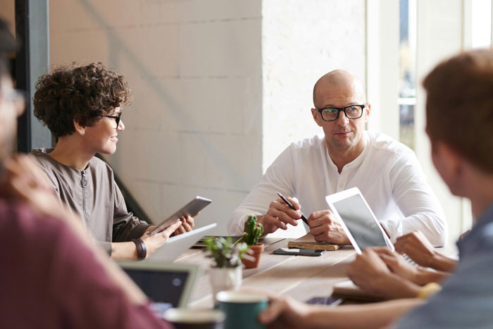 Manager discussing employee concerns with team, holding pen, with others using tablets in a modern office meeting room.