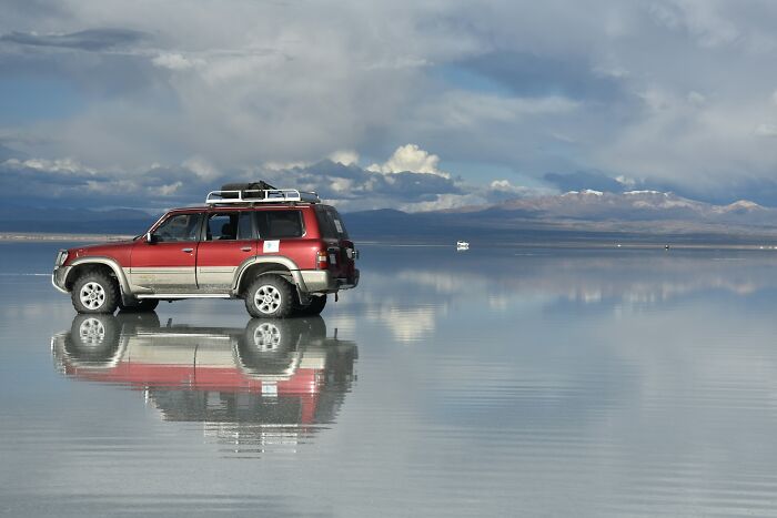 Red SUV parked on a reflective salt flat under dramatic skies, showcasing one of the wonders that prove the world is still full of magic.