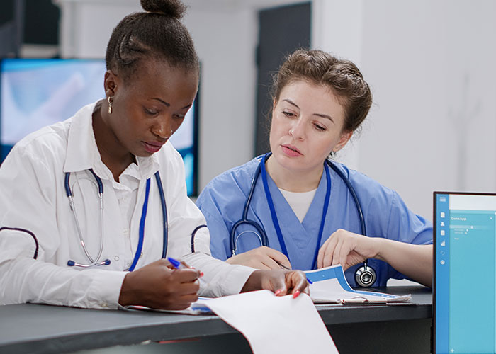 Two medical workers in scrubs and lab coats reviewing documents together in a busy medical facility.