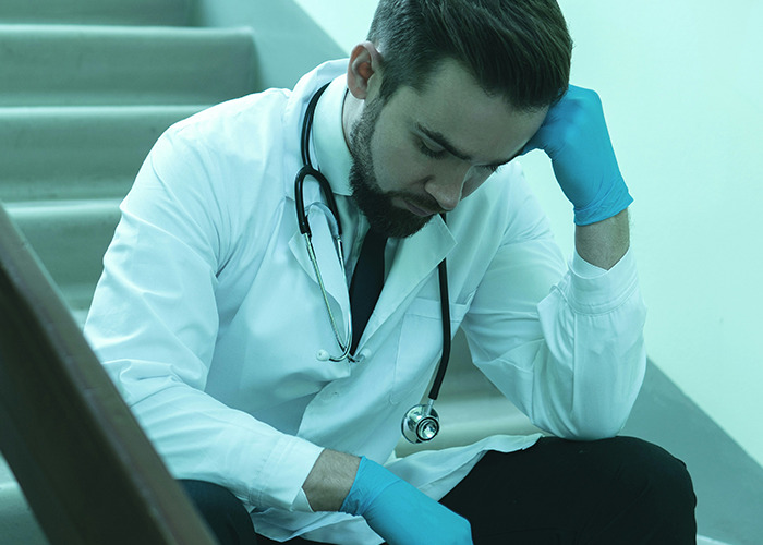 Male medical worker wearing a lab coat and blue gloves sitting on stairs looking stressed while pretending to stay calm