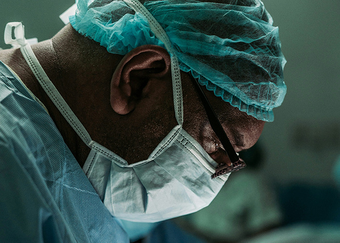 A medical worker wearing surgical cap, mask, and glasses, maintaining calm focus during a critical moment in the operating room.