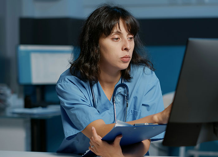 Female doctor in blue scrubs reviewing patient records on a computer, highlighting hurtful doctor behaviors.