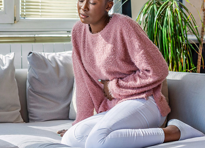 Woman in pink sweater sitting on couch, holding stomach in discomfort, reflecting hurtful doctor patient experience.