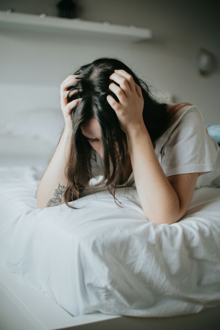 Woman sitting on bed holding head in hands, illustrating stress and symptoms of mundane things that make people sick.