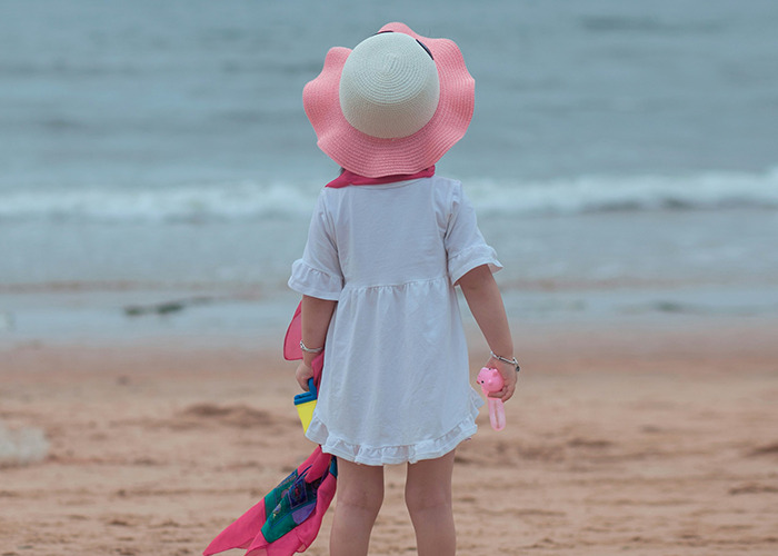 Child wearing a sun hat and white dress standing on the beach near the water, reflecting calm and peaceful vibes.