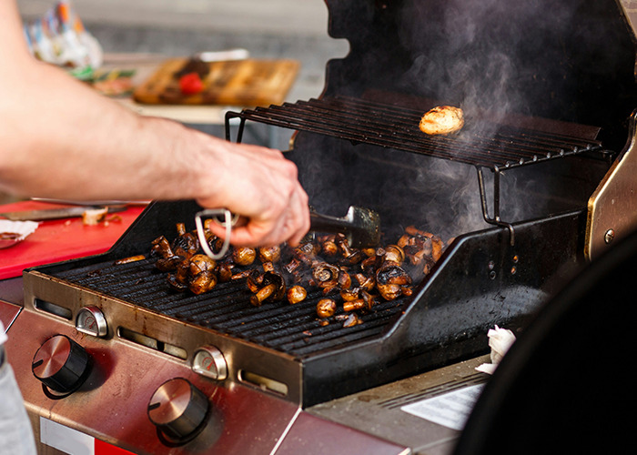 Close-up of mushrooms grilling on a barbecue with smoke rising, person turning them with tongs outdoors cooking.