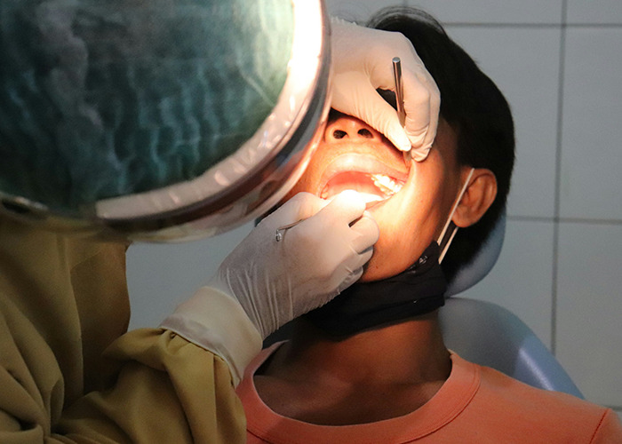 Medical field employee wearing gloves examines patient's teeth with dental tools under bright light in clinic setting.