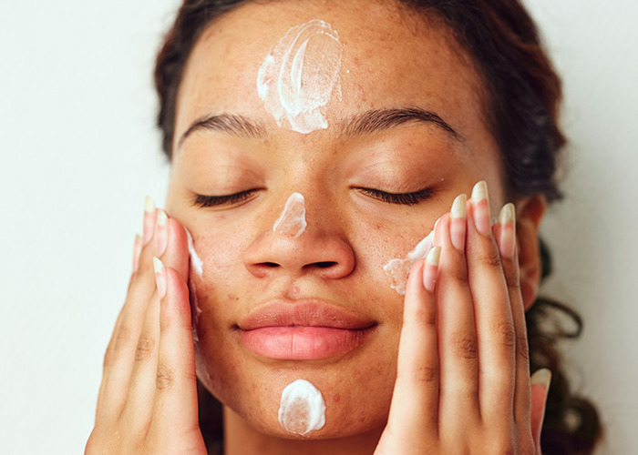Young woman with closed eyes applying cream on face, representing medical field employees caring for skin health.