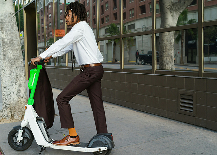 Medical field employee dressed formally riding an electric scooter on a city sidewalk near office buildings.