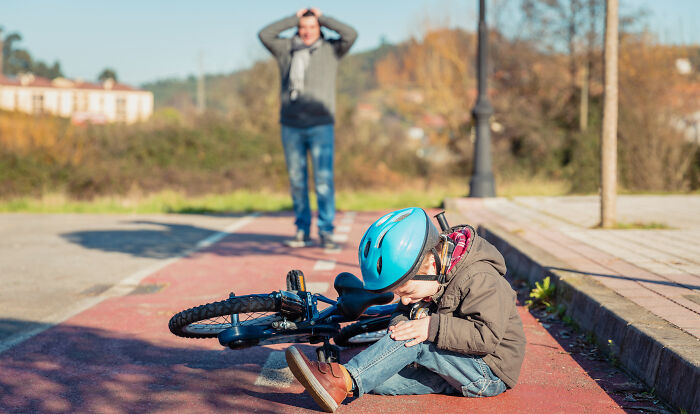 Child wearing a blue helmet sitting on the ground with a bicycle, highlighting unique body features after a fall.