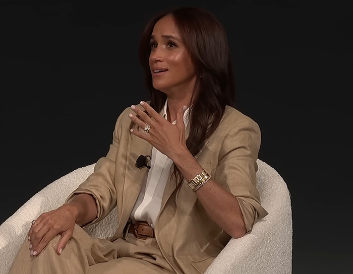 Meghan Markle speaking during an interview, wearing a beige suit, related to wedding caterer and Queen Elizabeth intervention.