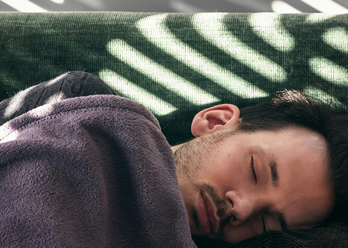 A man sleeping peacefully on a green couch wrapped in a blanket, illustrating moments men experience differently.