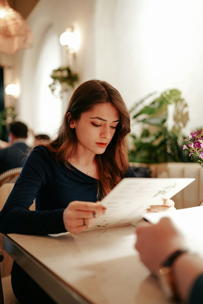 Young woman in a dark top focused on reading a menu at a bright restaurant, illustrating turn off behaviors men notice.