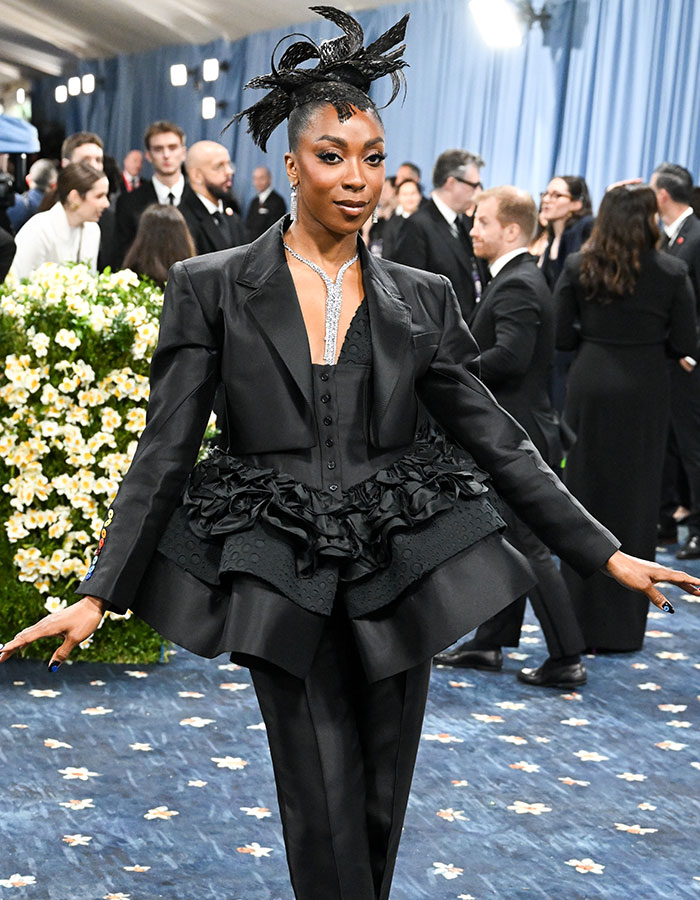 Stylish attendee in layered black outfit posing on the Met Gala 2025 red carpet with floral backdrop and crowd behind.