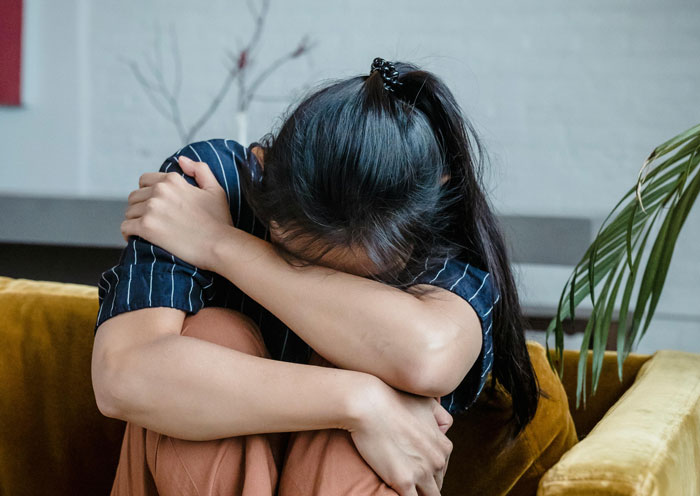 Young woman hugging knees while sitting on couch, feeling overwhelmed as mil calls herself mommy in tense moment