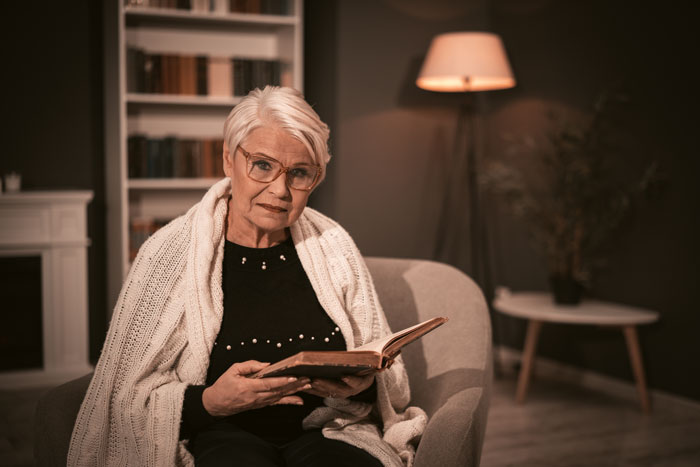 Older woman sitting in armchair at home, wrapped in blanket, holding book with concerned expression in dimly lit room. Older woman sitting in armchair at home, wrapped in blanket, holding book with concerned expression in dimly lit room.