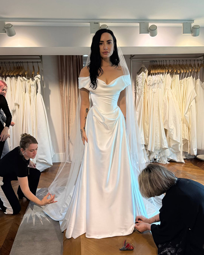 Woman wearing a white wedding dress and veil, with two stylists adjusting the gown in a bridal boutique.
