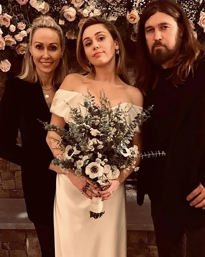 Miley Cyrus wearing off-shoulder wedding dress holding a bouquet, posing with two people against a floral backdrop.