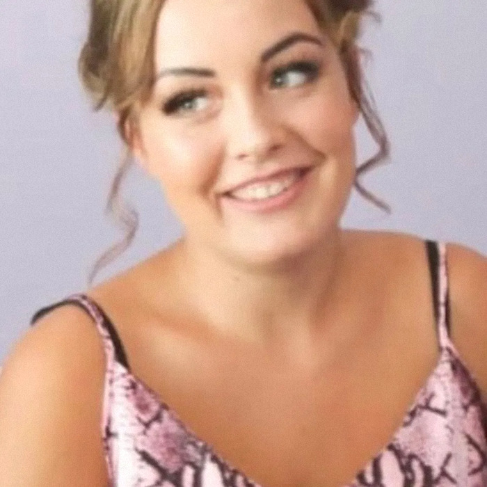 Young woman smiling with styled hair and wearing a pink patterned dress in a casual indoor setting.