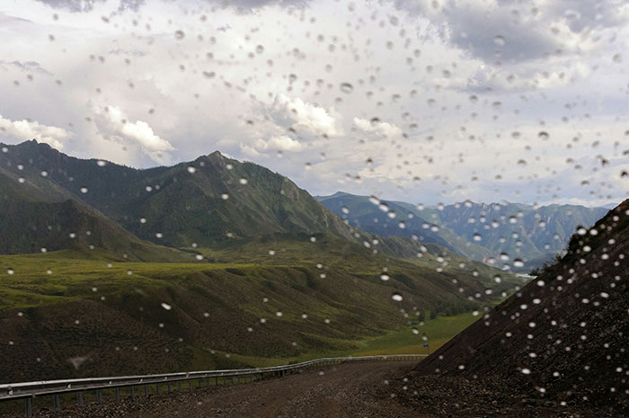 Mountain landscape viewed through a rain-speckled window with a winding road, evoking instinct and caution in nature.