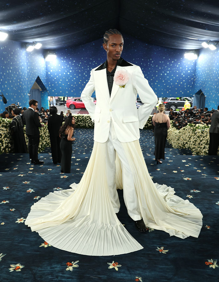 A model wearing a white suit with a long pleated train and a flower detail on the red carpet at Met Gala 2025.