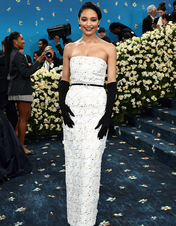 Actress in white floral gown and black gloves posing on the Met Gala 2025 red carpet with floral decorations in background.