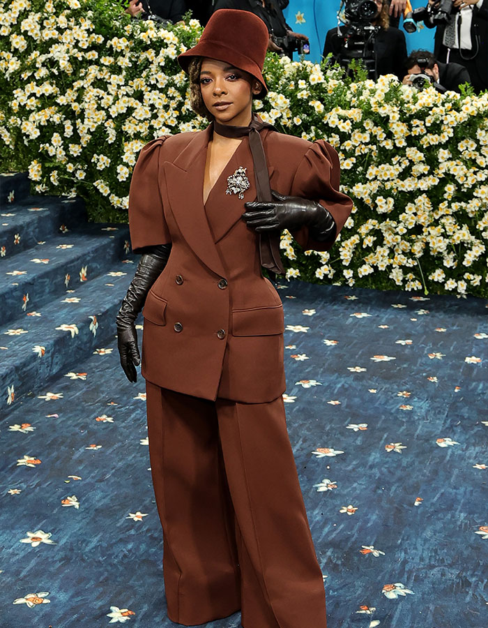 Woman in brown wide-leg suit and hat with black gloves posing on floral carpet at Met Gala 2025 red carpet event.