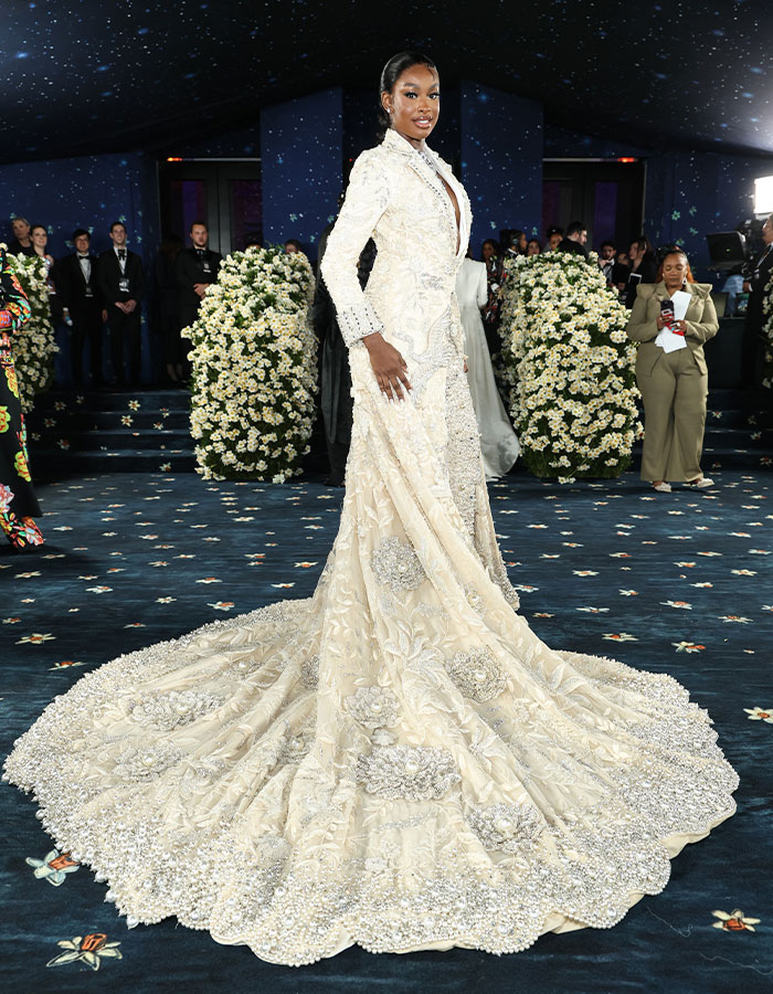 Woman in an intricate cream gown posing on the red carpet at Met Gala 2025 amid floral decorations and attendees.
