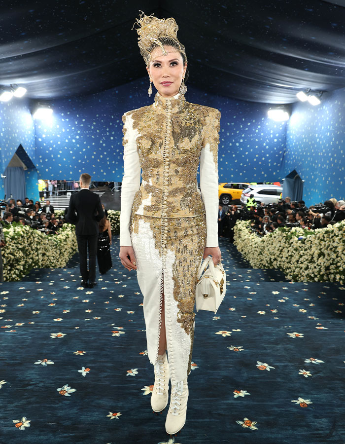Woman in gold and white embellished outfit and headpiece posing on Met Gala 2025 red carpet with photographers and floral decor behind.
