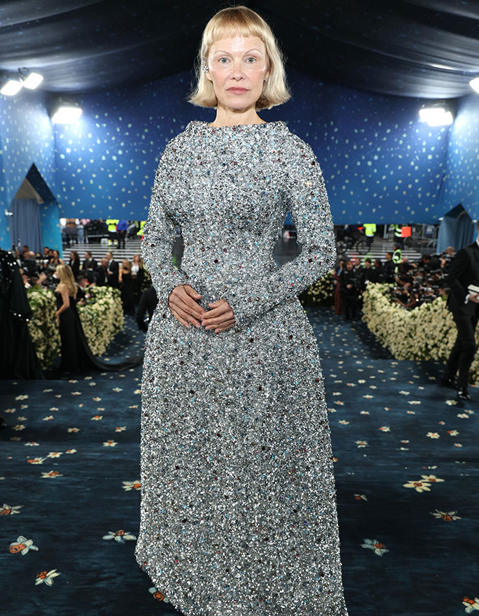 Woman in a sparkling silver gown posing on the Met Gala 2025 red carpet with photographers and floral decor in the background