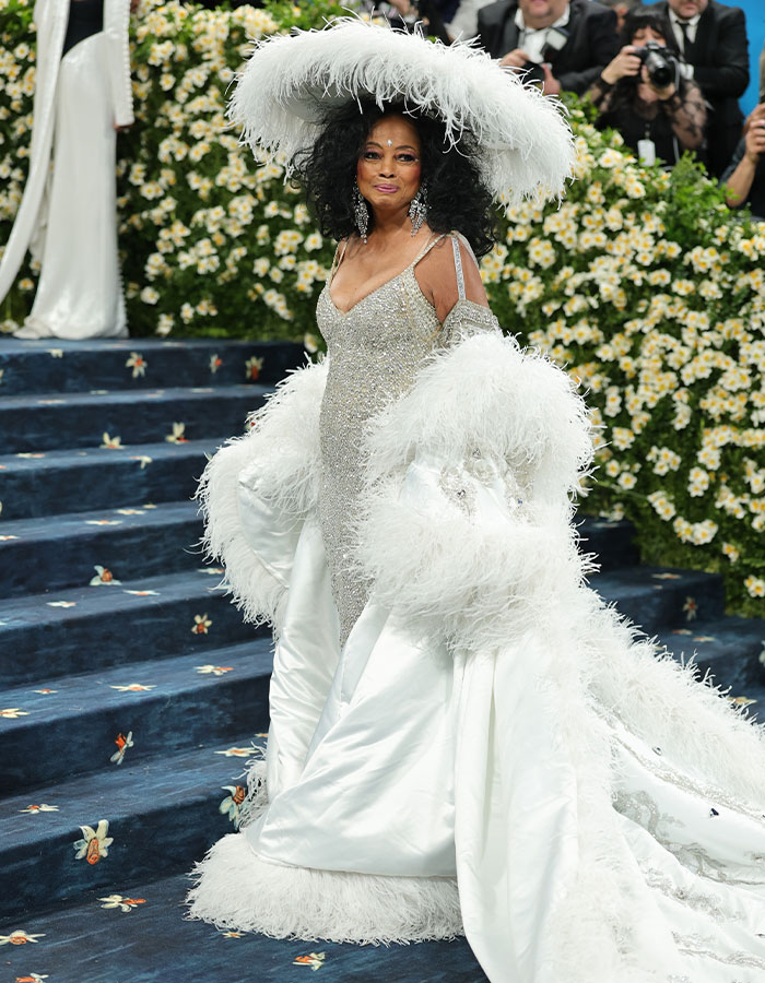 Woman in an elaborate white feathered gown and hat posing on the Met Gala 2025 red carpet with photographers in the background