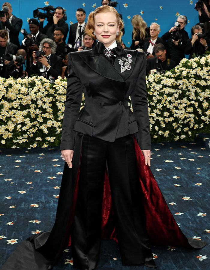 Actress wearing a black tailored suit with red lining on the Met Gala 2025 red carpet, photographers in the background.