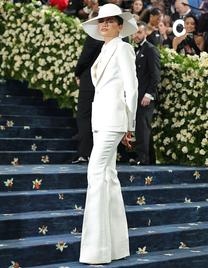 A woman in a white suit and wide-brimmed hat posing on the Met Gala 2025 red carpet with floral decorations.