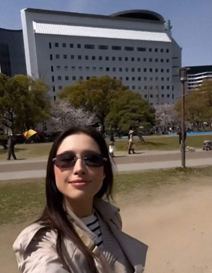 Woman solo traveler wearing sunglasses and coat taking a selfie outdoors with trees and a large building in the background