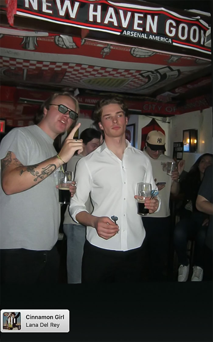 Two men holding drinks in a dimly lit room, capturing massive thirst over guy in Lana Del Rey Met Gala photo.