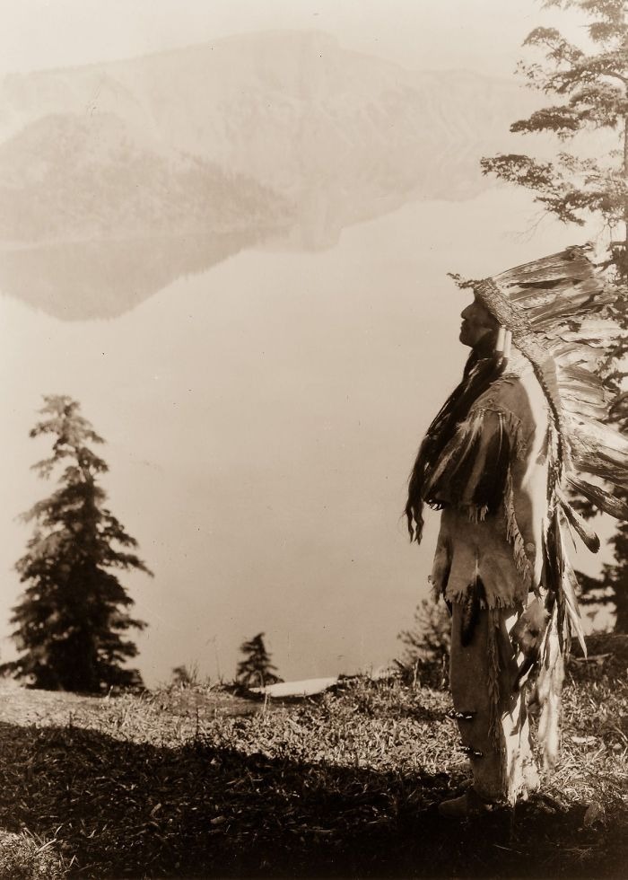Native American man in traditional headdress standing near a lake, captured in a powerful portrait from over a century ago.