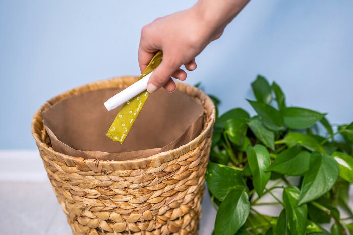 Hand discarding a wrapped item into a woven trash bin next to a green plant, hinting at company secret revealed.