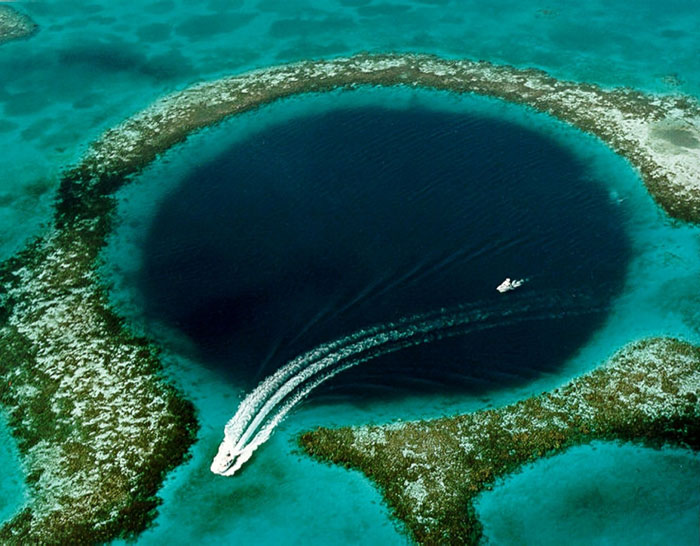 Aerial view of a rare natural phenomenon with boats cruising near a deep blue circular sinkhole in clear turquoise water.