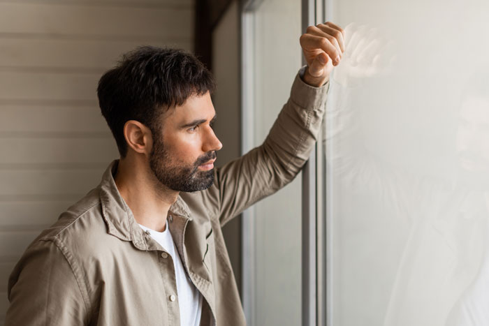 Man with beard looking thoughtfully out window, symbolizing teen parks on neighbor's property conflict and his mom’s defense. Man with beard looking thoughtfully out window, symbolizing teen parks on neighbor's property conflict and his mom’s defense.