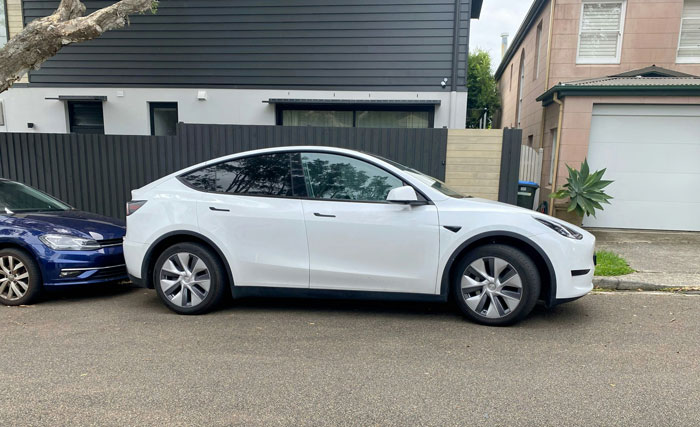 White electric car parked on neighbor's property with a blue car behind it in a residential street setting. White electric car parked on neighbor's property with a blue car behind it in a residential street setting.