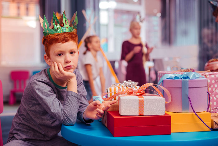 Sad boy wearing a crown sitting by birthday presents, reflecting exclusion and family conflict at a child's party.