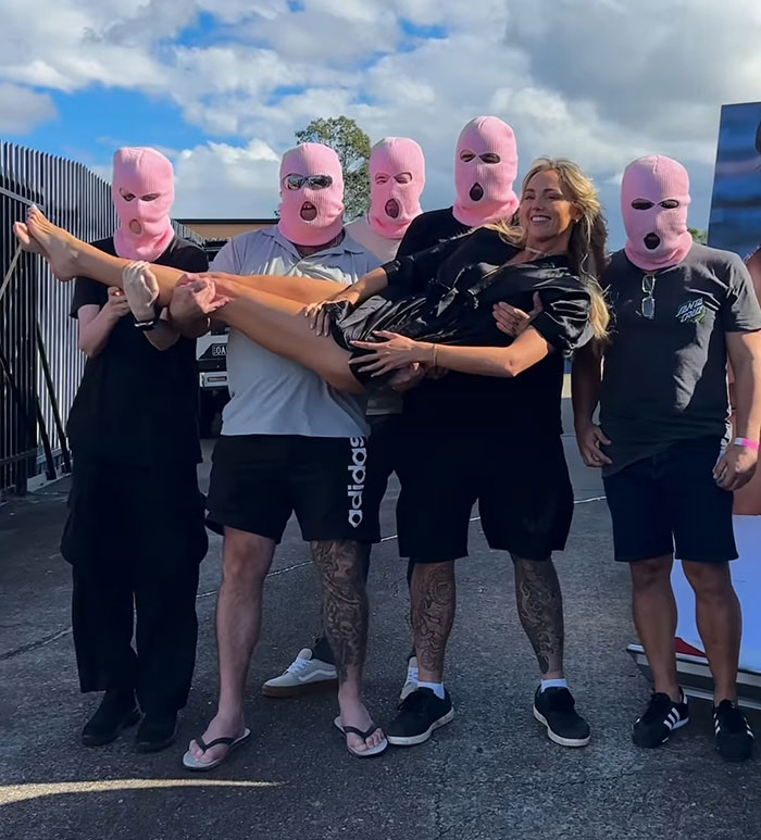 Annie Knight posing outdoors smiling while being held by five men wearing pink ski masks on a sunny day. Annie Knight posing outdoors smiling while being held by five men wearing pink ski masks on a sunny day.