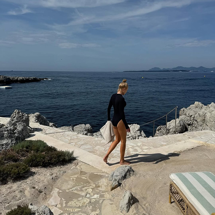 Woman with natural hair in a black swimsuit walking near rocky shore under clear sky, reflecting Nicole Kidman wig fail moment.