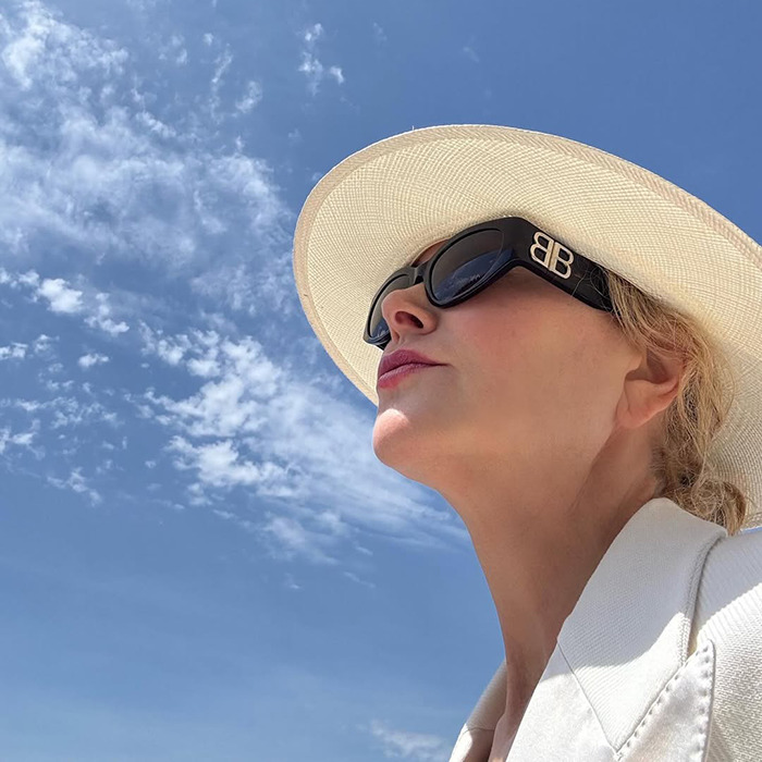 Nicole Kidman outdoors wearing black sunglasses and a white hat, showing her natural hair under a clear blue sky.