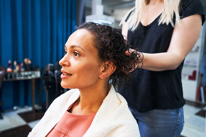 Woman with alopecia sitting in salon chair while stylist works on her hair, highlighting alopecia and headscarf issues.