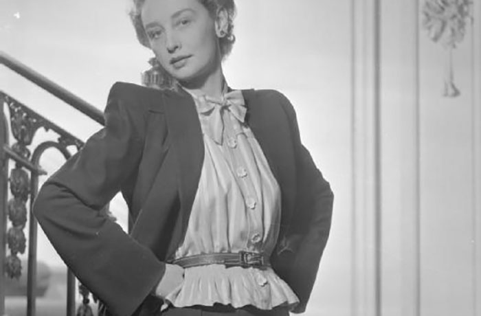 Black and white image of a woman posing confidently wearing a blazer and blouse, reflecting vintage stirrup pants fashion trends.