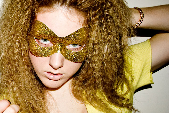 Young woman with crimped hair wearing a gold glitter mask and yellow top, styling inspired by vintage fashion trends.