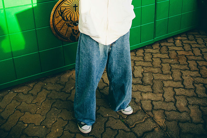 Person wearing wide denim jeans and sneakers standing on a brick floor near green tiled wall featuring stirrup pants style.